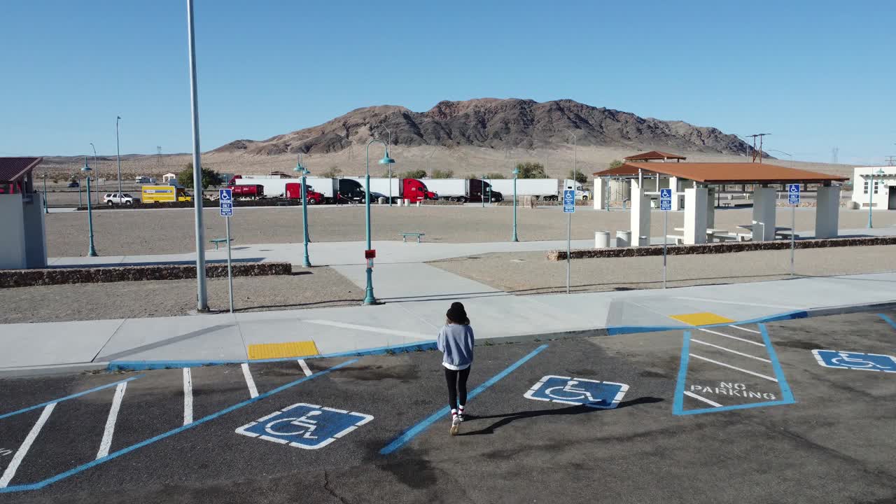 Faceless woman walking through quiet highway rest stop surrounded by desert terrain, clear sky, distant mountain range and parked trucks in West Virginia