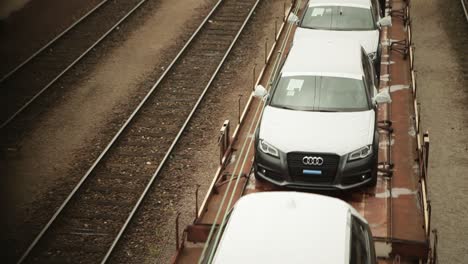 Static-camera-shot-of-a-train,-carrying-new,-white-cars-along-a-twin-line-brown-railway-in-an-industrial-setting