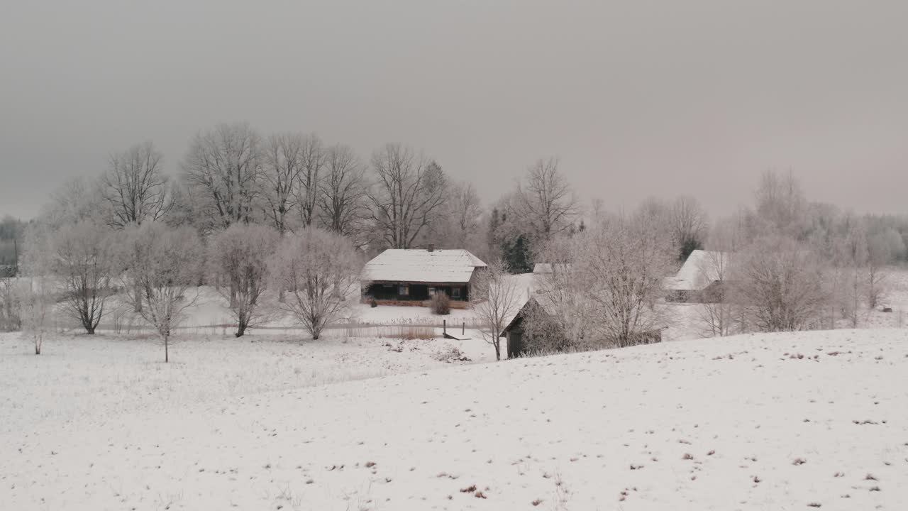 Wide drone shot of a historic wooden family home with bath house and barn in countryside landscape. Winter season with lots of snow and frost on the trees on a cloudy day.