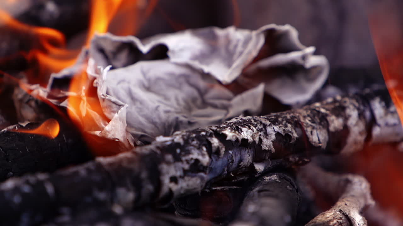Fire tongues over the smouldering logs. Paper twisting in fire. Embers of burnt-out wooden logs in slight flame. Close-up. Slow motion.