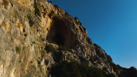aerial view flying close to a wall, toward a cave carved in a mountain, in greece