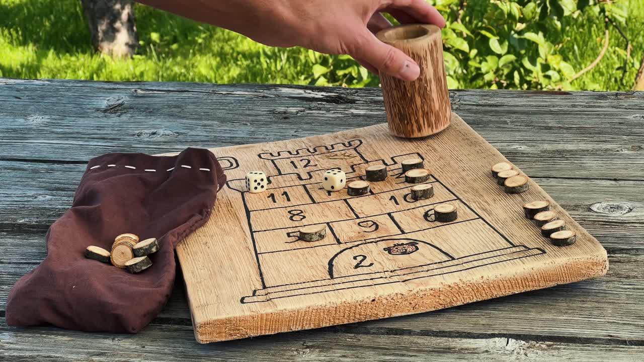 Hand moving a small wooden pawn on a handmade wooden board game with dice, a dice cup, and a bag of pawns, suggesting a fun and engaging activity