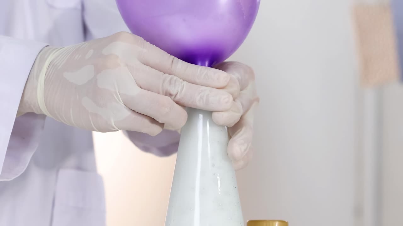 A purple balloon inflates as a white liquid reacts inside a bottle, handled by gloved hands.