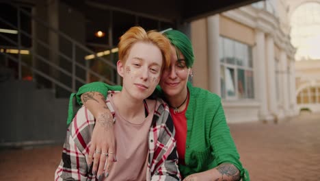 retrato: dos chicas lesbianas felices en su cita. una chica rubia con un corte de pelo corto y chispas en su cara en una camisa rosada a cuadros y su novia con cabello corto verde en una camiseta verde miran a la cámara con una mirada coqueteante