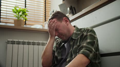 A-sad-thoughtful-and-despairing-brunette-man-with-stubble-in-a-checkered-shirt-holds-his-head-while-he-thinks-about-his-life-while-sitting-on-the-floor-in-the-kitchen-during-the-day