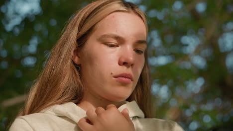 close-up of lady in casual hoodie holding her hood collar, intently focused on phone, sunlight gently illuminates her face through leaves, casting soft shadows, with blurred greenery in background