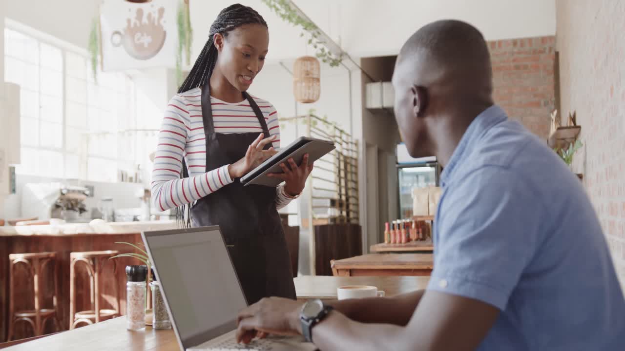 African american female barista taking order on tablet from male customer using laptop, slow motion
