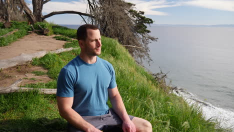 a man sitting in a meditation pose after a workout to practice mindfulness and stress relief on a beach cliff over the blue ocean in santa barbara, california