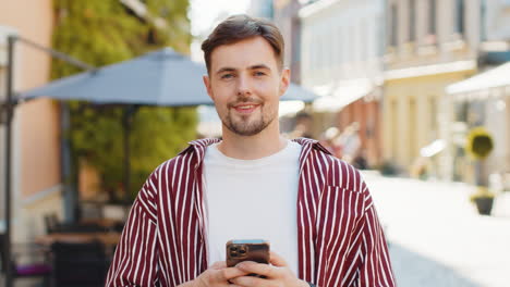 un hombre con una camisa a rayas sonriendo y sosteniendo su teléfono mientras camina por una calle.