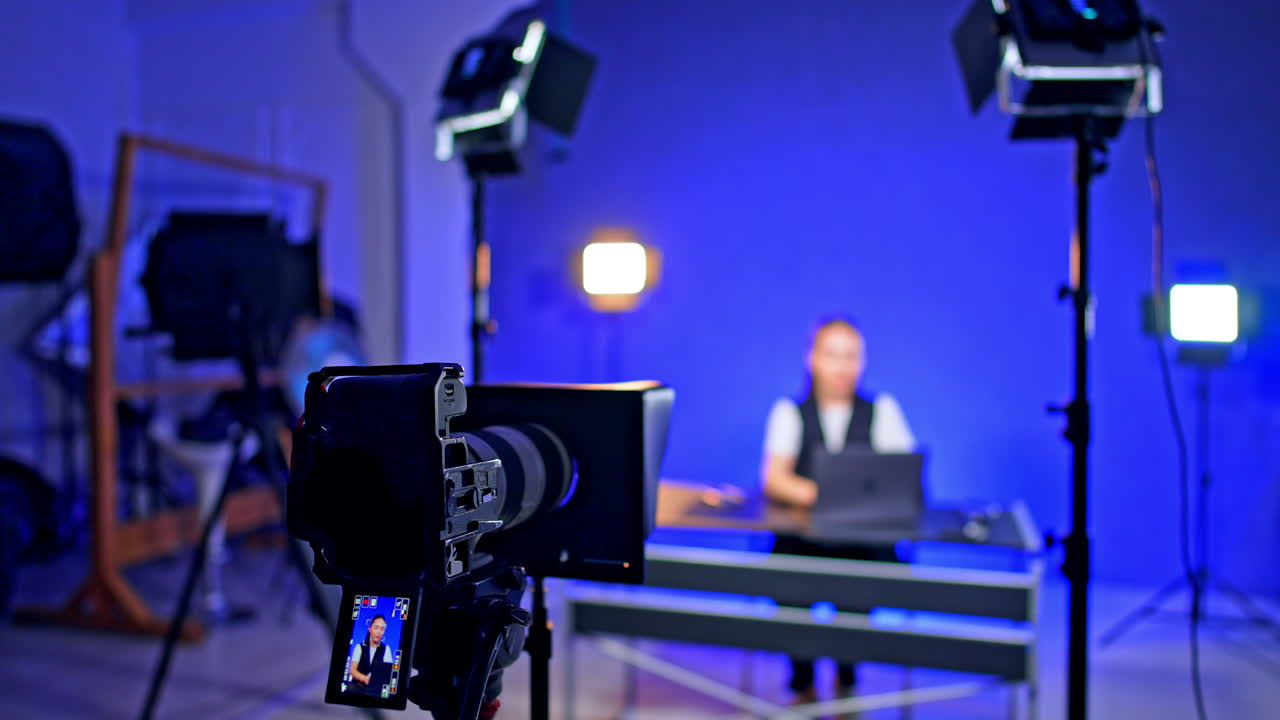 Studio video setup. A person sits at a desk with a laptop in a well-lit studio, preparing for a video recording session