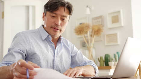 mature asian man sitting at table at home using laptop to organise household bills and finances - shot in slow motion