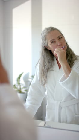 vertical video: brushing teeth, woman in bathrobe looking in bathroom mirror in morning routine