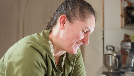 housewife in green dress and apron bends forward with soft, joyful smile in warm kitchen environment, sunlight highlighting content expression, while kitchen utensils and appliances blur