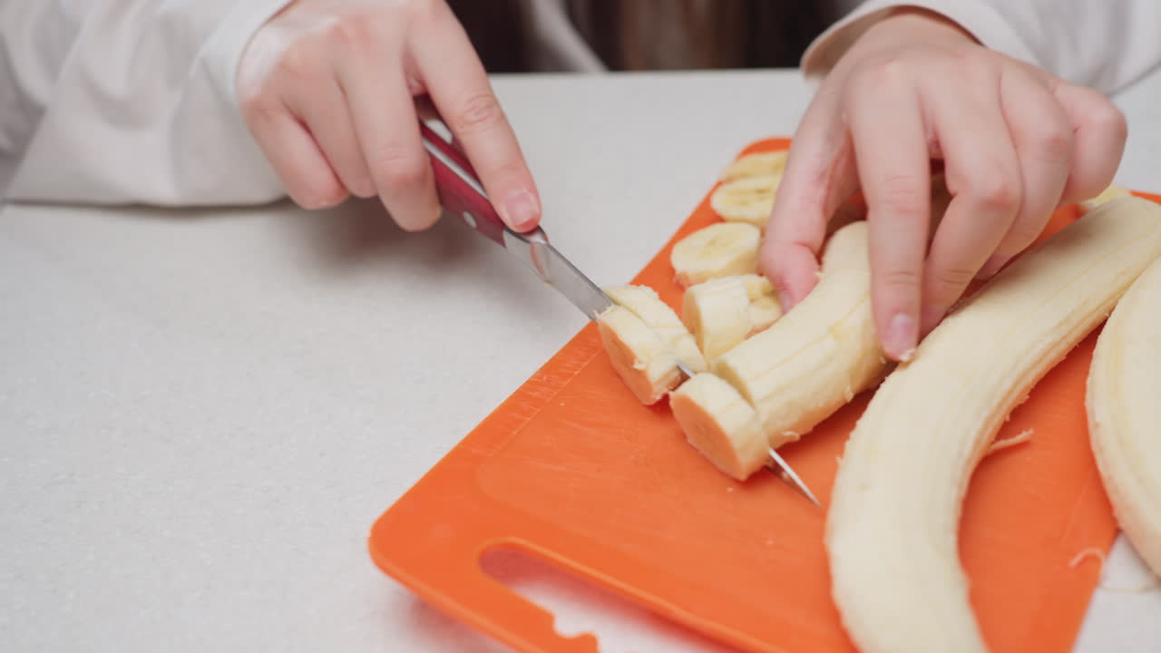 Overhead view female hands cutting banana into round slices on orange board, kitchen counter bright and clean, careful motion with knife, close focus on rings, fresh snack preparation
