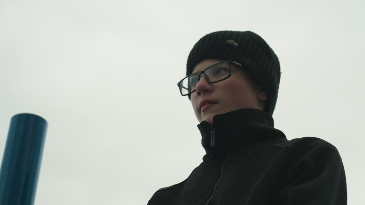 Close-up of a young boy in a black outfit and glasses, standing near an iron bar, looking down adjusted his glasses