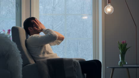 man in sweater feeling stressed in a cozy room as snow falls outside, hand on head, pensive mood, ambient lighting