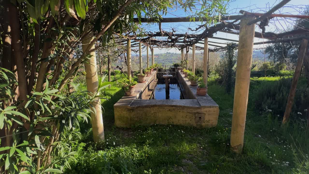 A beautiful, long stone fountain under a rustic pergola in a sunlit garden, surrounded by plants and overlooking the rolling hills of Umbria, Italy