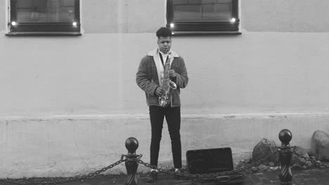 Blanco-Y-Negro-Y-Vista-Frontal-De-Un-Estuche-De-Saxofón-En-El-Suelo-Y-Un-Hombre-Con-Chaqueta-Tocando-El-Saxofón-En-La-Calle