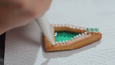 close-up of cookie decoration in progress with white icing on green-colored filling, three green beads placed at edge, creating intricate design on white tissue paper background