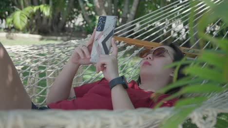 static shot of a girl relaxing on a hammock while using a phone during a sunny and windy morning