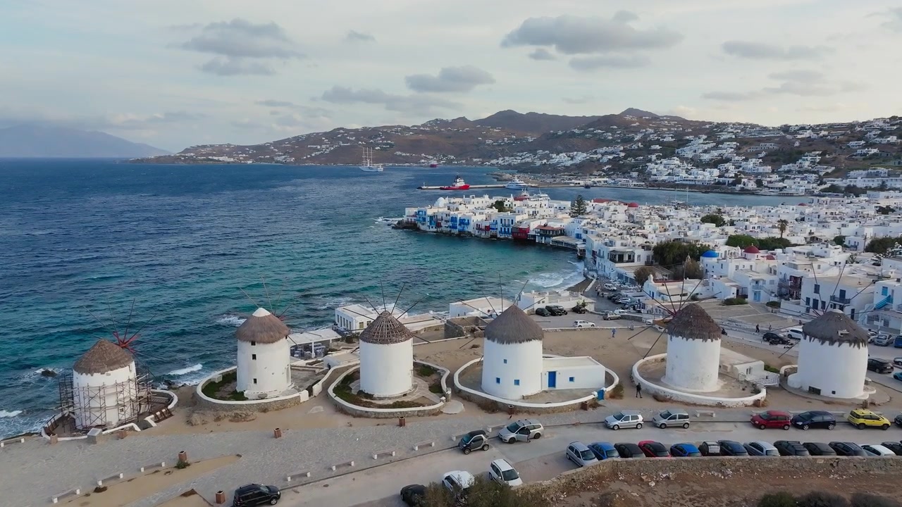 Yunanistan, Kiklad Adaları'nda gün doğumu sırasında Mykonos yel değirmenlerinin üzerinden Küçük Venedik'e doğru yavaş drone uçuşu. Dağınık bulutlar, altın rengi sabah ışığı.