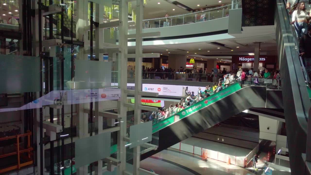 People on an escalator in a busy shopping mall with glass walls and multiple floors in daylight