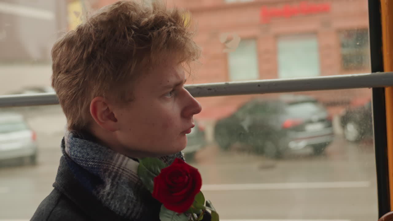 Young white creator holding red rose, on tram window seat, scarf bundled, city street passing in rain, pensive profile gaze, hands clutching stem, cinematic winter light, romantic solitude, creator