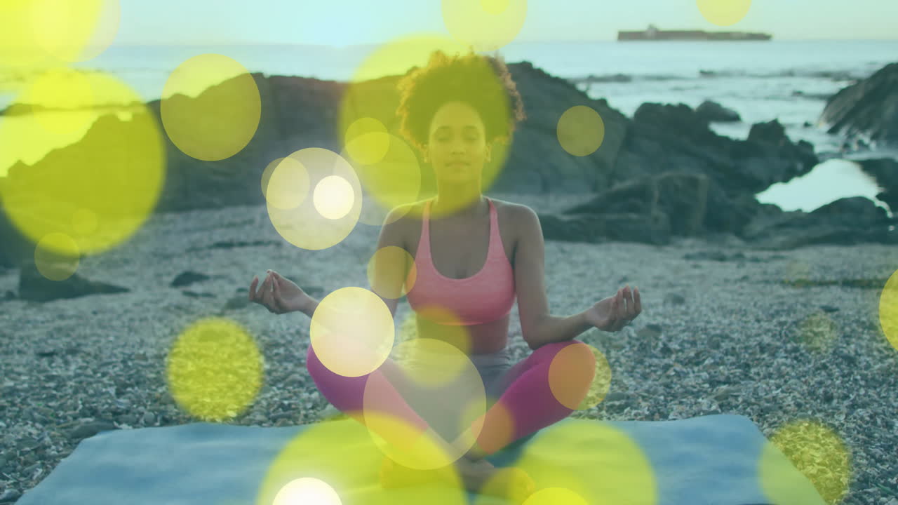 woman meditating on blue yoga mat at pebble beach, showing wellness with floating bokeh lights