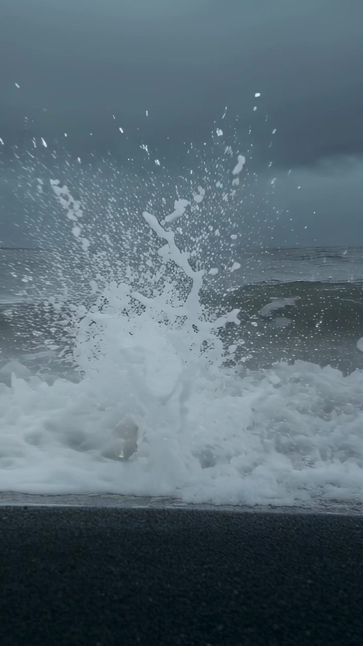 Vertical video: Rolling incoming swell collapsing spraying white foam receding on black sand shore
