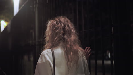 Woman in white coat walking with bag touching iron fence during peaceful night stroll with soft blurred background showing people walking down sidewalk and parked cars illuminated by streetlights