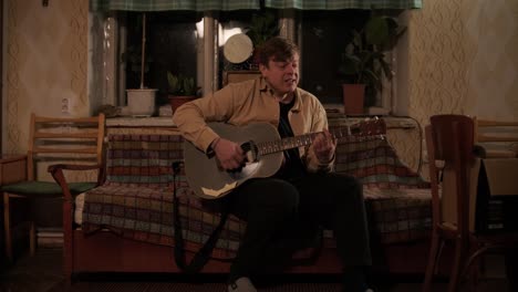 pretty young man playing guitar while sitting on sofa in dark living room