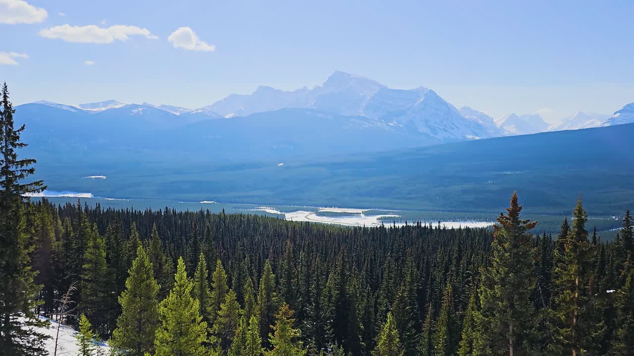 Vista panoramica di montagne innevate che si ergono sopra una fitta foresta di conifere e luminosi canali fluviali, mostrando una vasta natura selvaggia, un'atmosfera frizzante e una bellezza naturale scenografica.