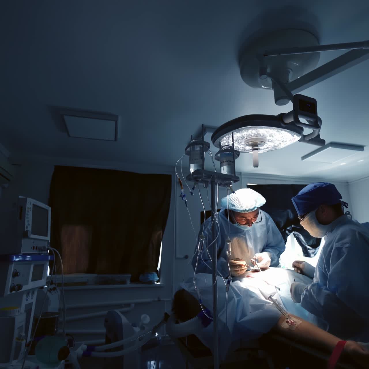 Surgery under the lamp in dark operating room. Professional doctors conduct an operation to a patient in the intensive care unit. Saving life in hospital.