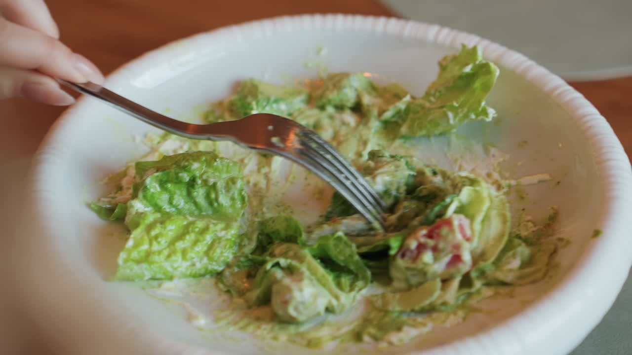 Inquadratura media di una forchetta che mescola insalata verde con avocado e pomodoro su un piatto di carta, pranzo da studente in un caffè del campus con movimento vivace e atmosfera informale, un boccone veloce tra le lezioni, luce naturale