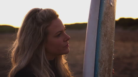 Frau-Im-Neoprenanzug-Hält-Surfbrett-In-Der-Hand-Und-Genießt-Den-Surfurlaub-Am-Strand,-Während-Die-Sonne-Untergeht