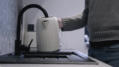 kitchen tap is running as man approaches it and places kettle under the flow so it fills up