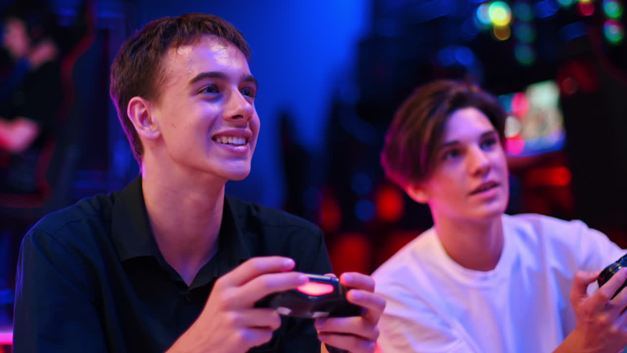 Dva dospívající kamarádi hrají na herní konzoli pomocí gamepadů, povídají si a usmívají se. Modré a červené osvětlení. Zpomalený záběr.
