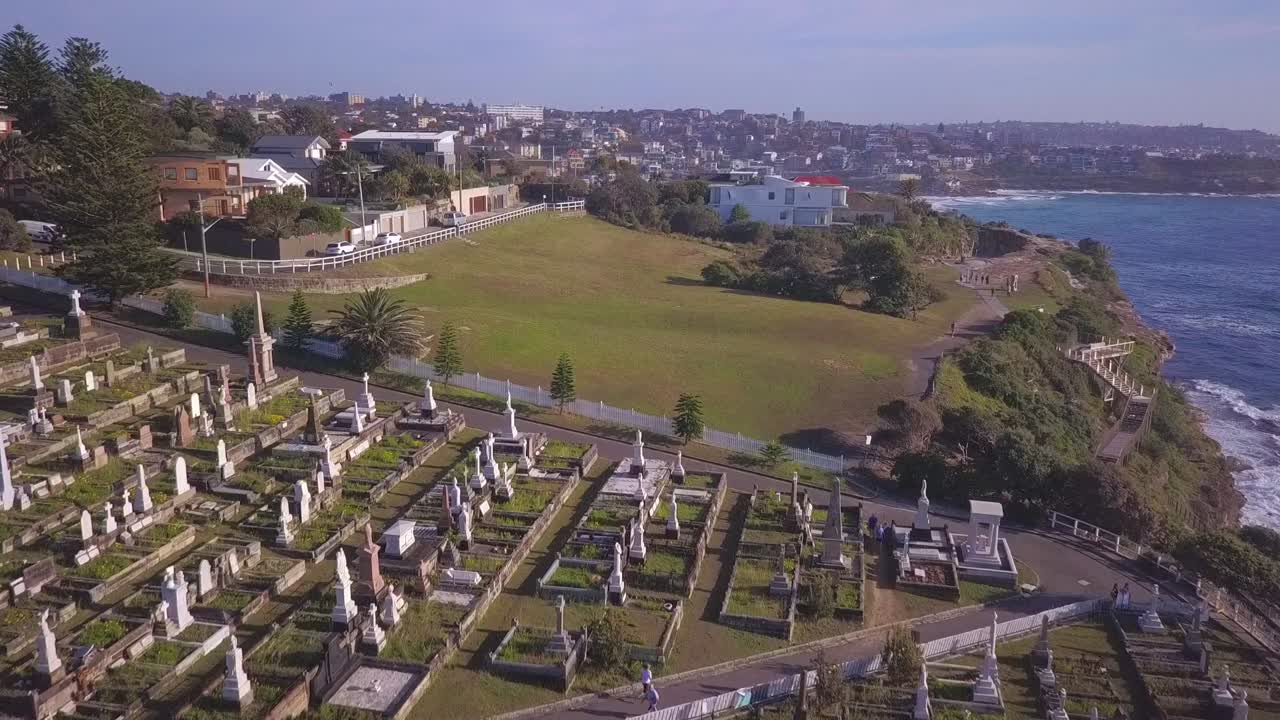 Aerial view over green cemetery on the cliff facing beautiful Pacific Ocean in Sydney Australia during the day under blue skies. Fly over top down bird eye view.