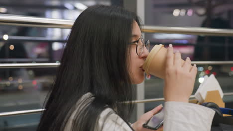 lady in white shirt places hand on wooden table while sipping coffee with calm and warm smile, seated in cozy chair near shiny metal railing and large glass window revealing blurred city night lights