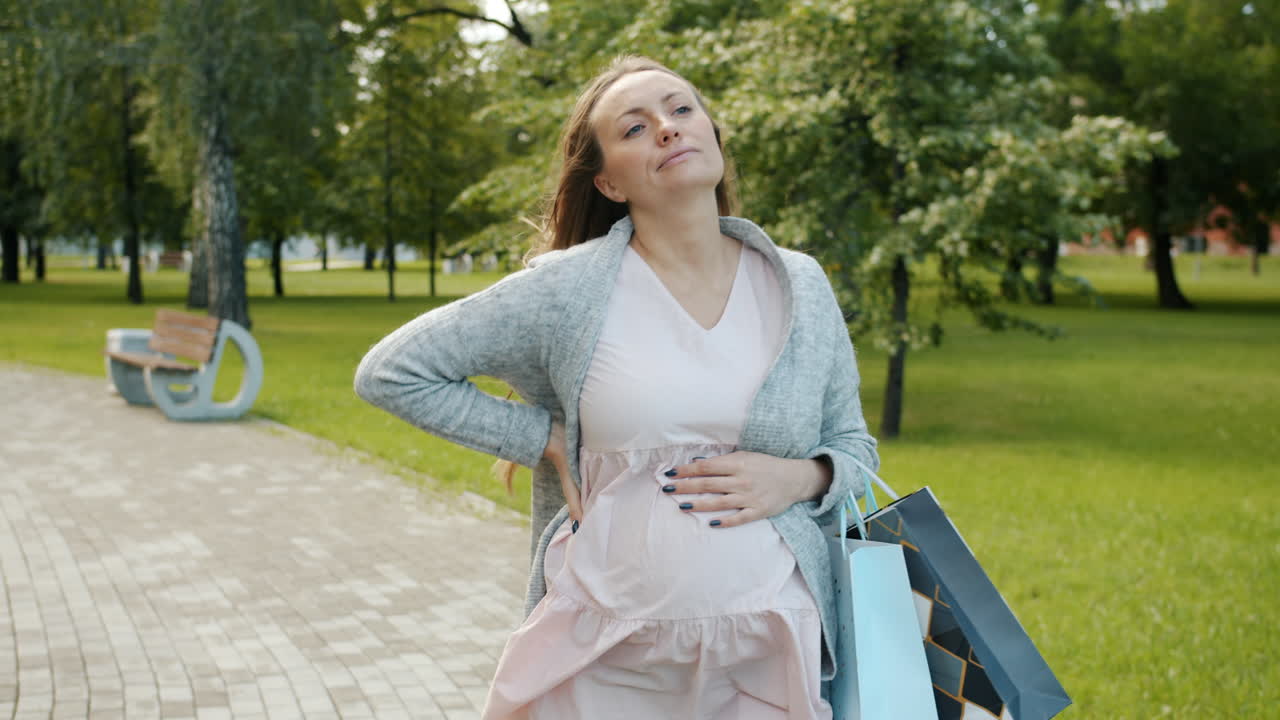 Pregnant Woman Walking in Park with Shopping Bags and Back Pain