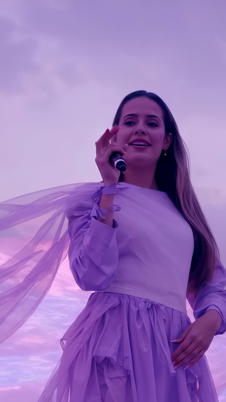 Vídeo vertical: cantor cantando contra o céu ao entardecer enquanto a brisa levanta um lenço transparente, com microfone sem fio
