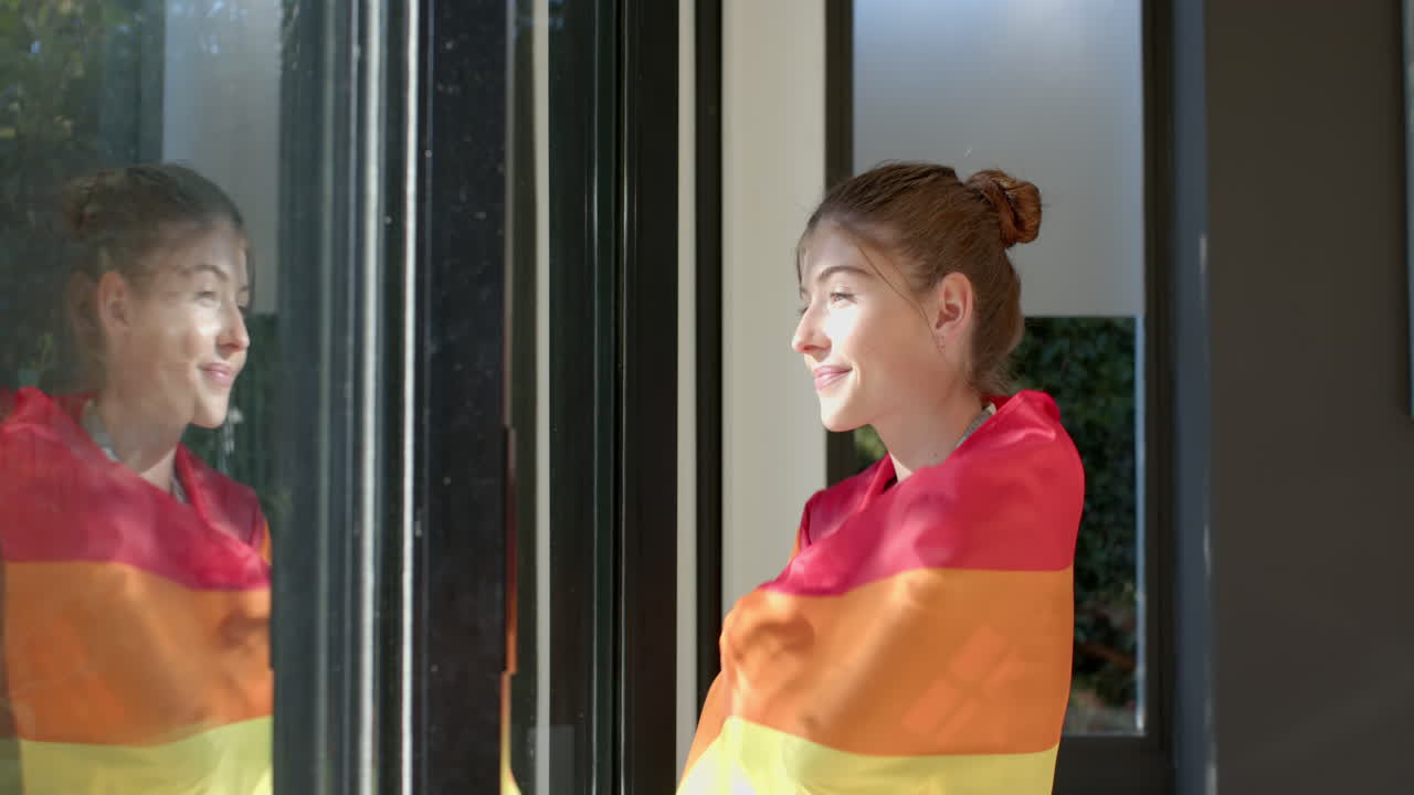 Smiling woman wrapped in rainbow flag looking out window, enjoying sunlight