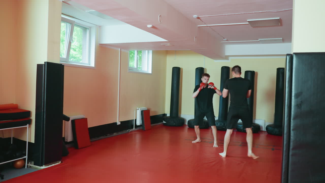 창문에서 들어오는 햇빛 아래, 붉은색 매트 바닥에서 서로 마주 보고 장갑을 낀 채 자세를 연습하며 샌드백으로 둘러싸인 무술 체육관에서 훈련 세션 동안 스파링하는 가라테 수련자