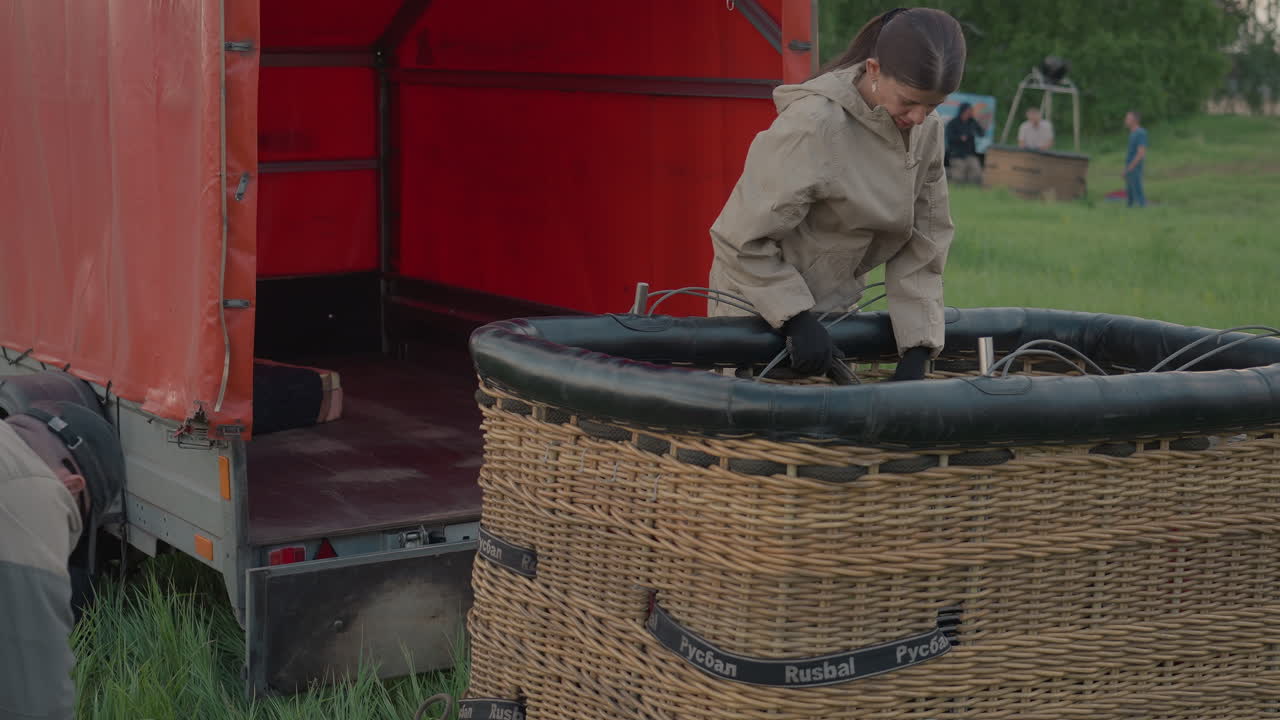 man and woman preparing hot air balloon wicker basket next to trailer in grassy field handling ropes and equipment for upcoming flight during calm morning light rural setting