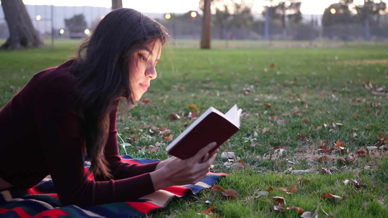 A hispanic girl reading a fictional story book or novel in the park at sunset