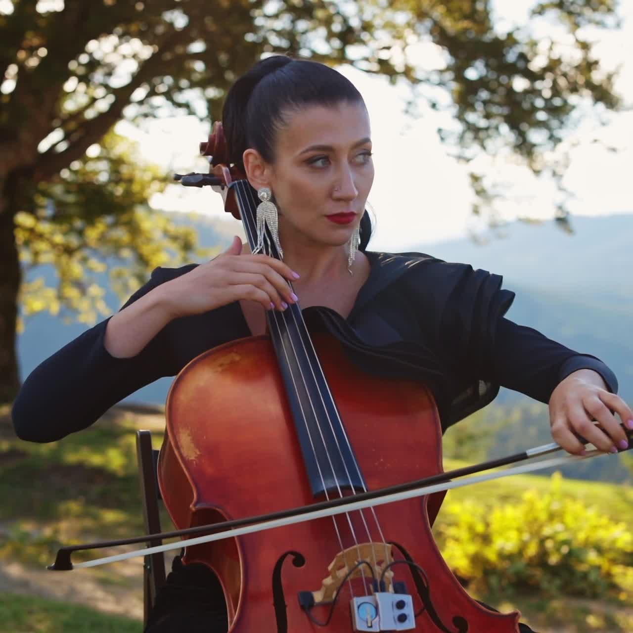 Violoncello musician with bright makeup and long earrings playing instrument. Lady stops to fix the earring and goes back to playing. Nature backdrop