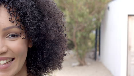 biracial woman with curly hair smiling warmly outdoors near modern home, copy space