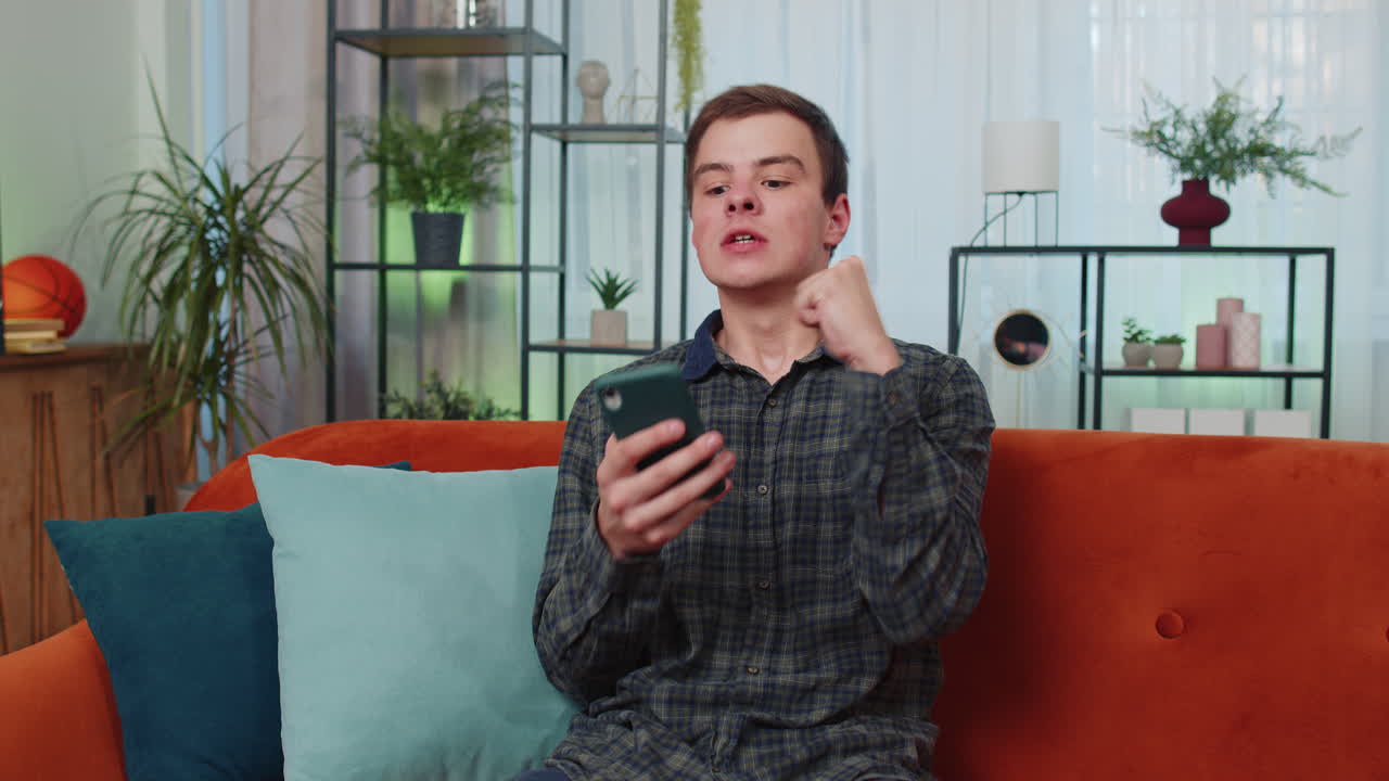 십대 젊은 남자가 집에서 스마트폰을 사용하여 승리를 축하합니다 복권 잭 ⁇  좋은 소식 큰 큰 승리