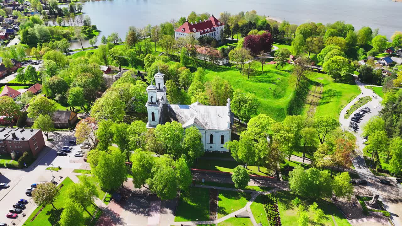 A white church and red-roofed estate rise from a lush riverside park, surrounded by winding paths, city buildings, and shimmering spring foliage in a European town.