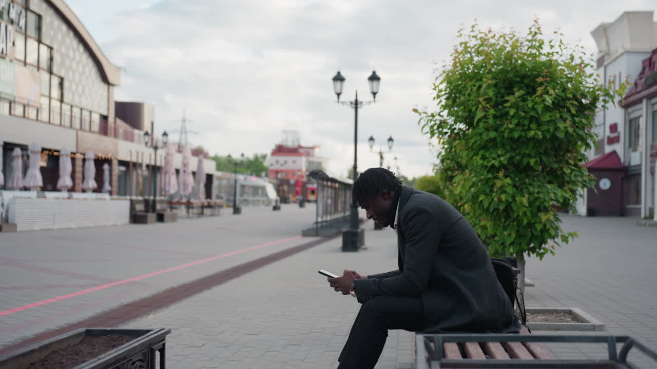 Business man walking towards bench on city street while using phone, holding briefcase, dressed in formal suit, showing modern urban lifestyle, focus, determination, and morning work routine
